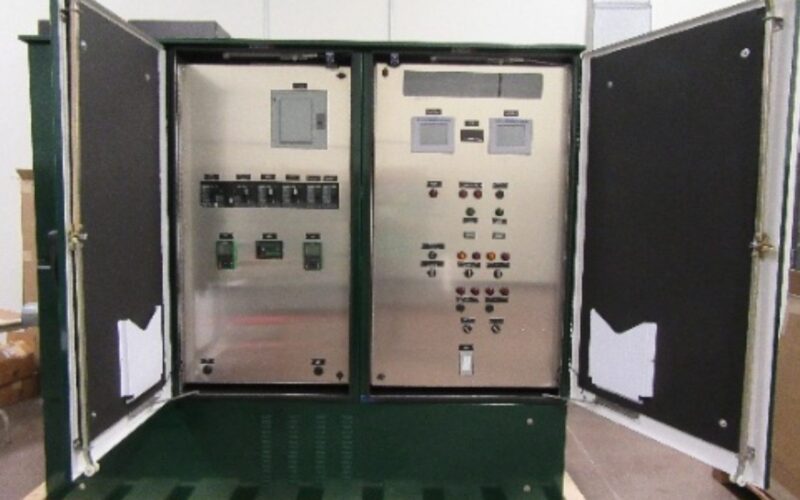 Screenshot 2026-05-26 at 1.58.56 PM A green electrical control panel cabinet with both doors open, revealing various switches, buttons, meters, and indicator lights on the control panels inside, situated on a wooden pallet indoors.