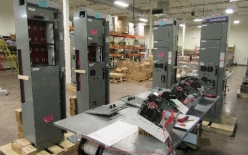 Screenshot 2026-05-26 at 1.59.06 PM Large electrical panels and switchboards are being assembled in a spacious industrial workshop, with various tools, wires, and boxes scattered around the workspace. Shelves and equipment are visible in the background.