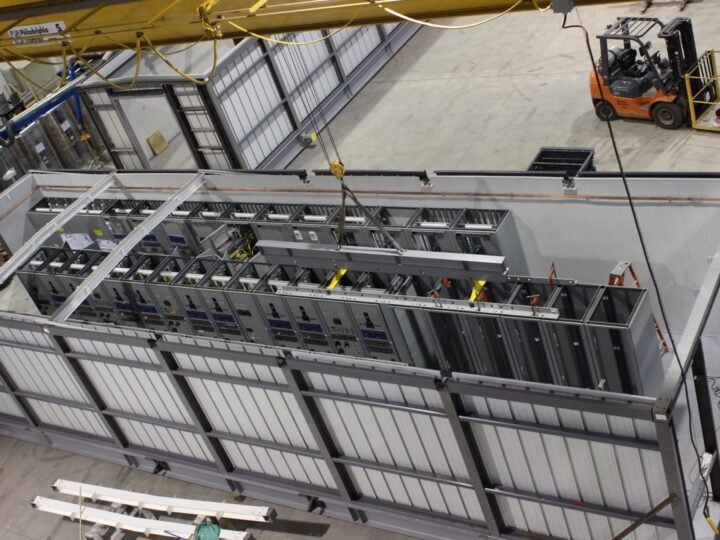 image009 A large industrial module under construction in a factory, with exposed metal framing, electrical panels, and equipment. A crane and a forklift are visible in the background, along with workers and ladders.